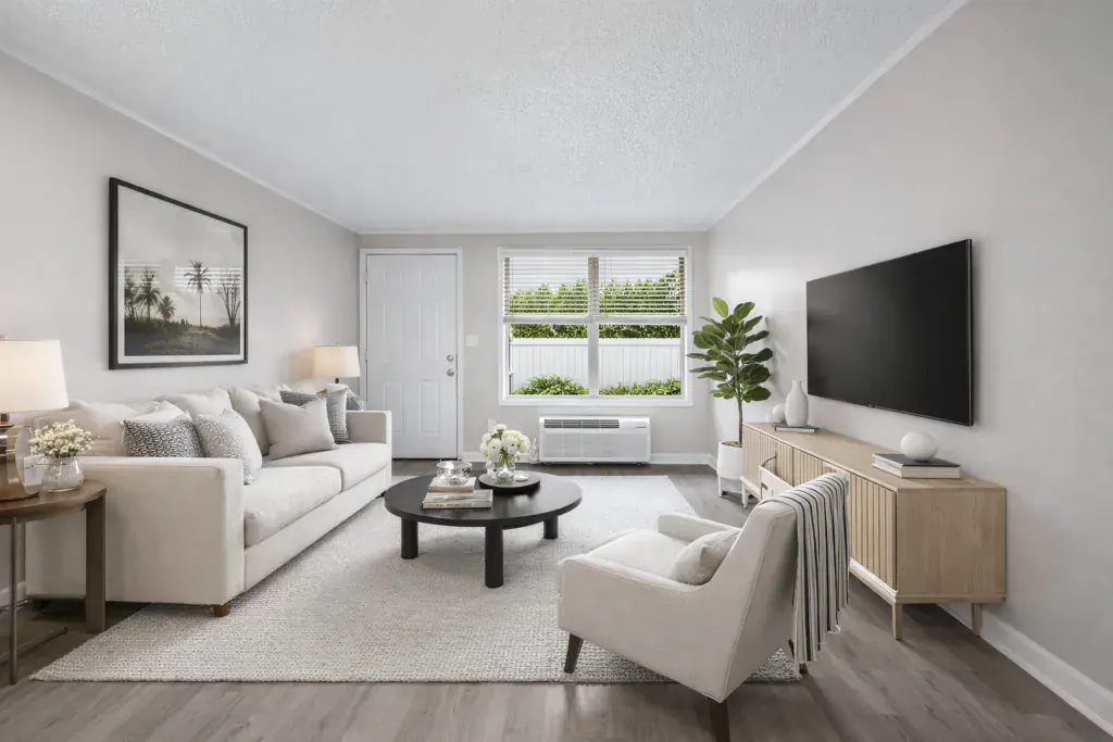 Furnished living room with a sofa and armchair beside a window looking onto greenery, with a through-wall air conditioning unit below