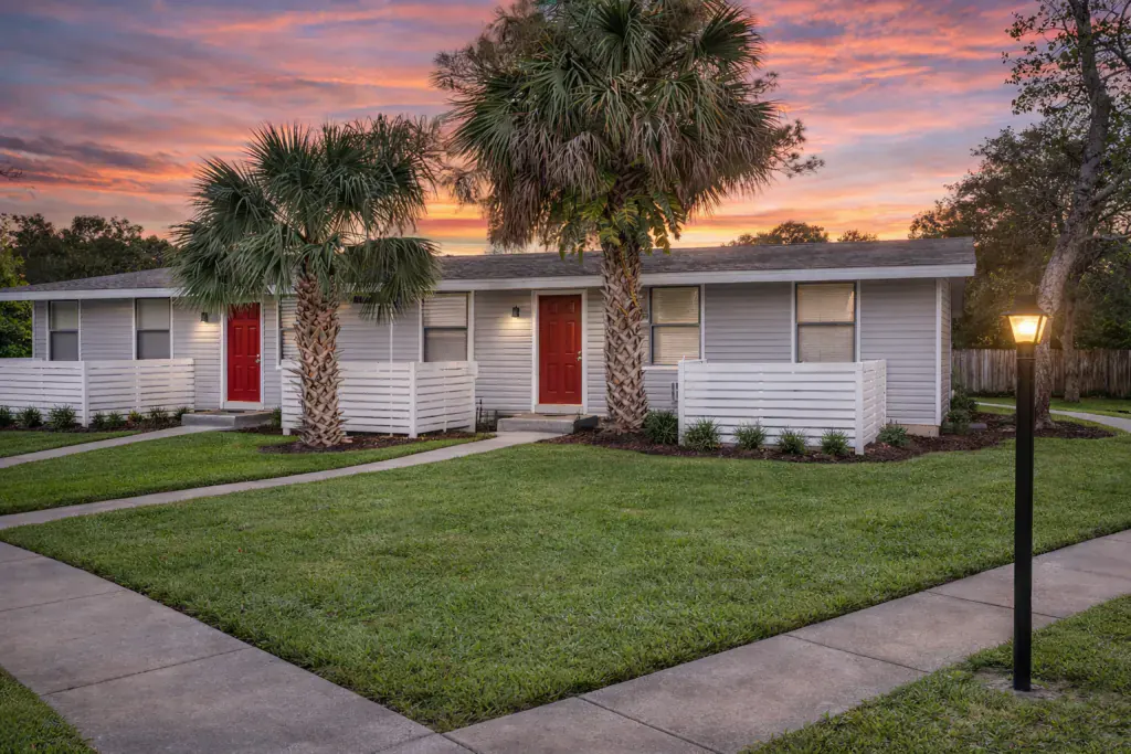Two single-story Lakewood apartments with red front doors and white slat privacy fences, palms and a lit lamp post on the lawn at sunset