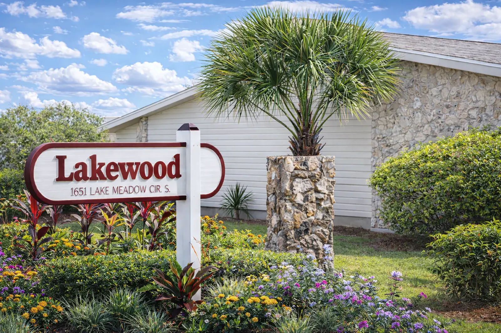 The Lakewood entrance sign reading 1651 Lake Meadow Cir. S., set in flowering beds beside a palm and a coquina stone column